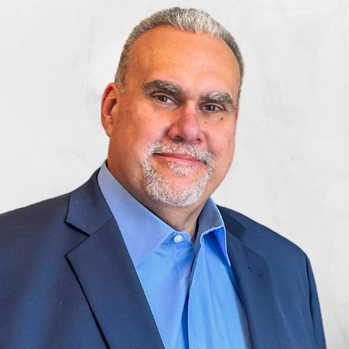 Man with short gray hair and goatee in a blue suit and shirt, smiling against a light background.