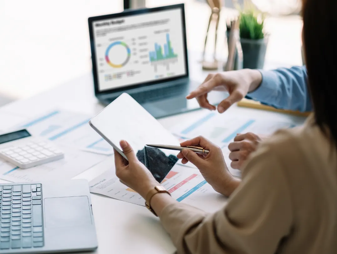 business-eval-1 Two people review data on a tablet and printed charts, with graphs displayed on a laptop in the background.