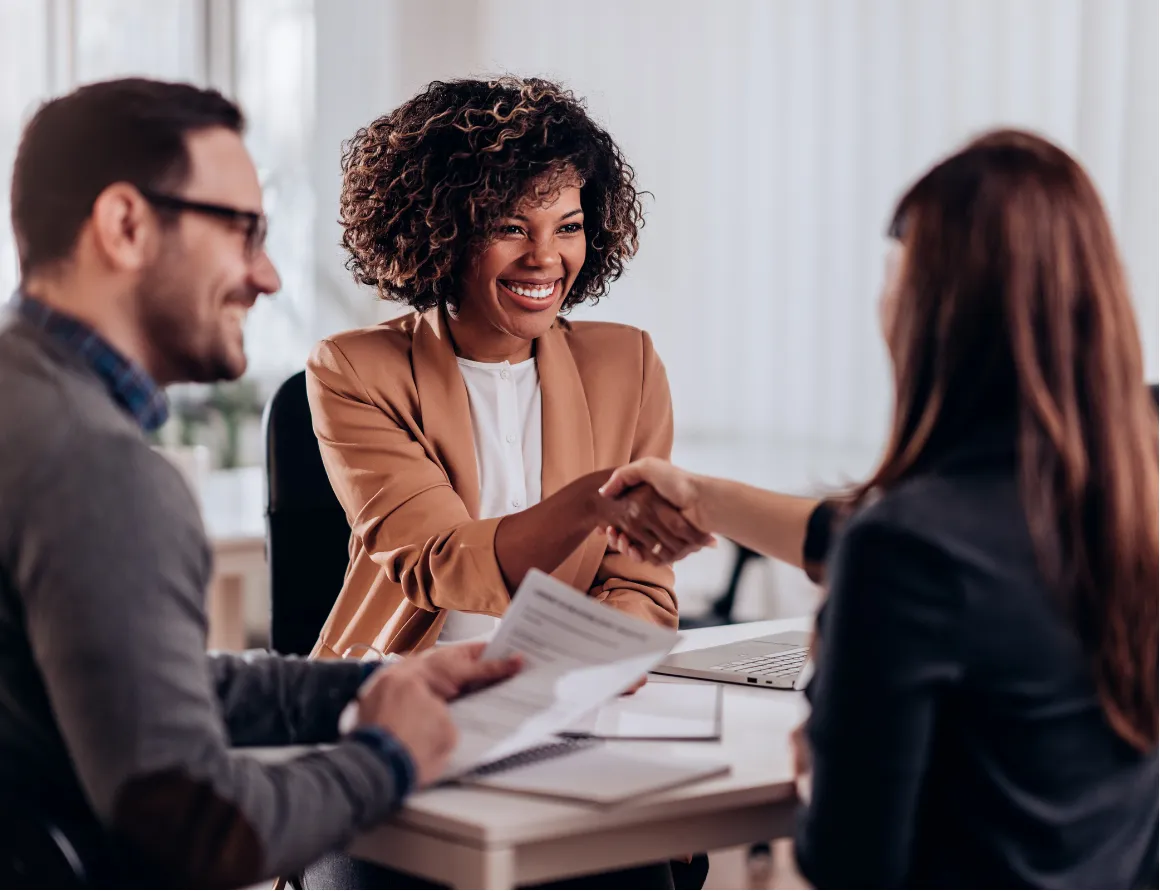 Three people in an office, two women shaking hands and a man holding documents, all smiling.