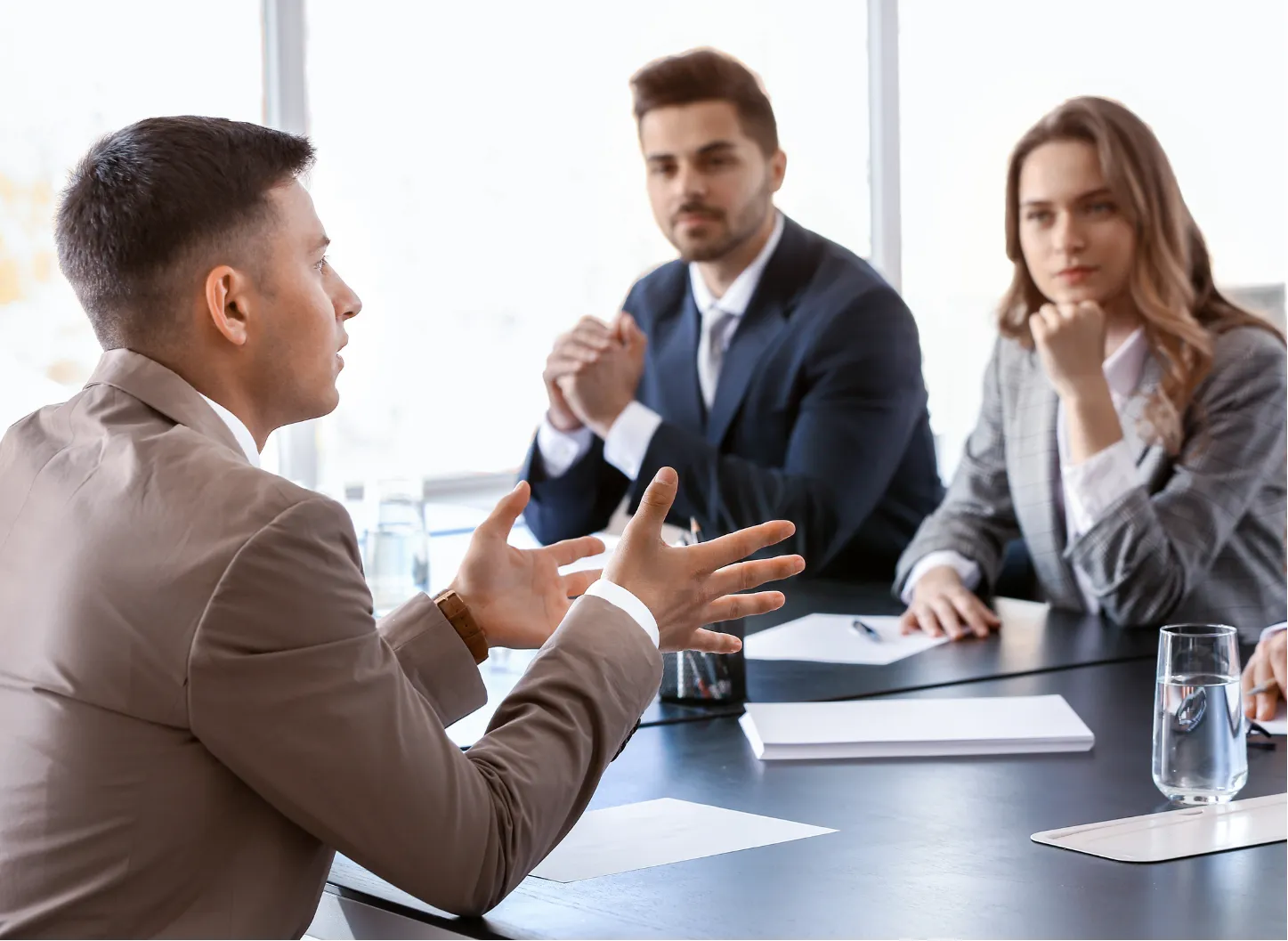 Three people in business attire listen to a man speaking at a meeting table with documents and glasses of water.