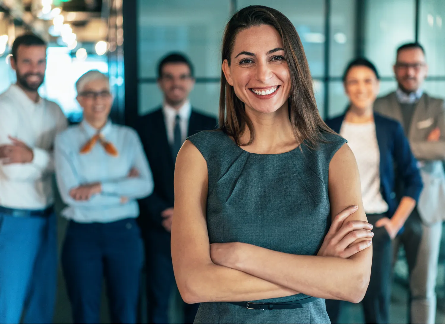 Smiling woman in business attire stands confidently with arms crossed, colleagues blurred in the background.