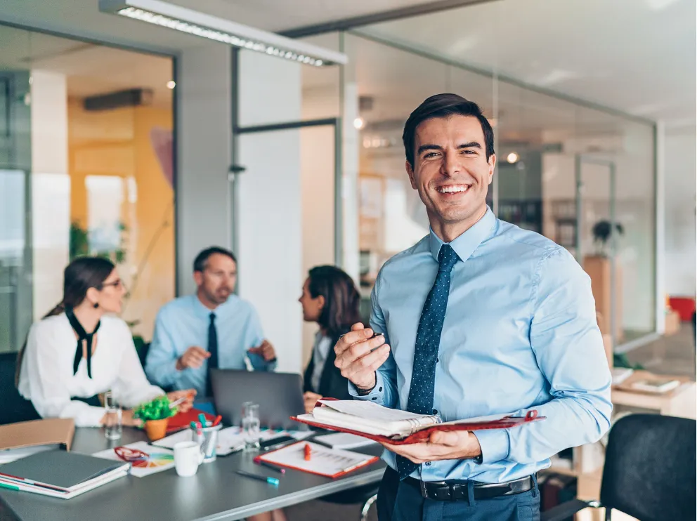 manage-1 Smiling man in business attire holding documents, with colleagues having a meeting in the background.