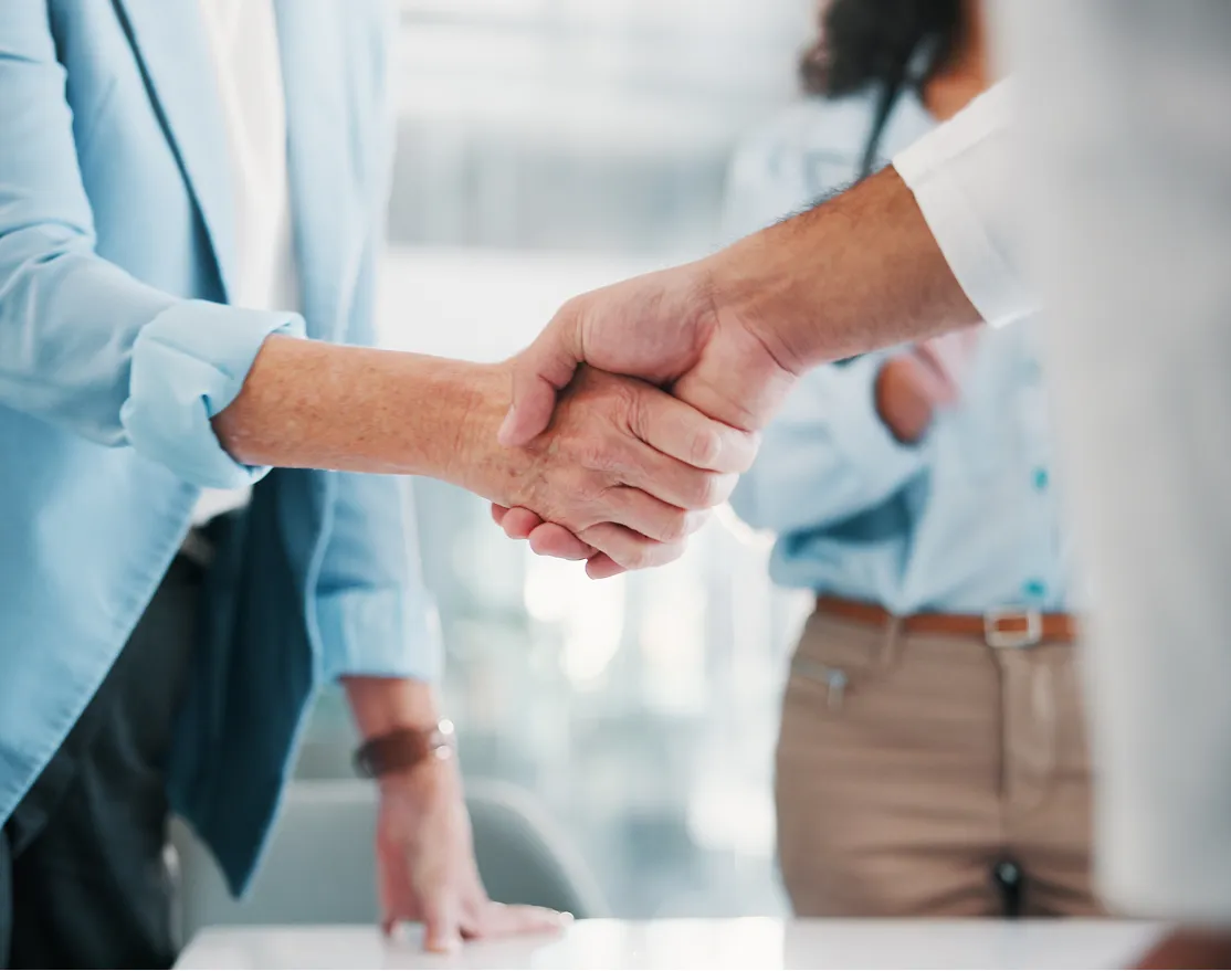 Two people in business attire shaking hands in an office setting, with a third person in the background.