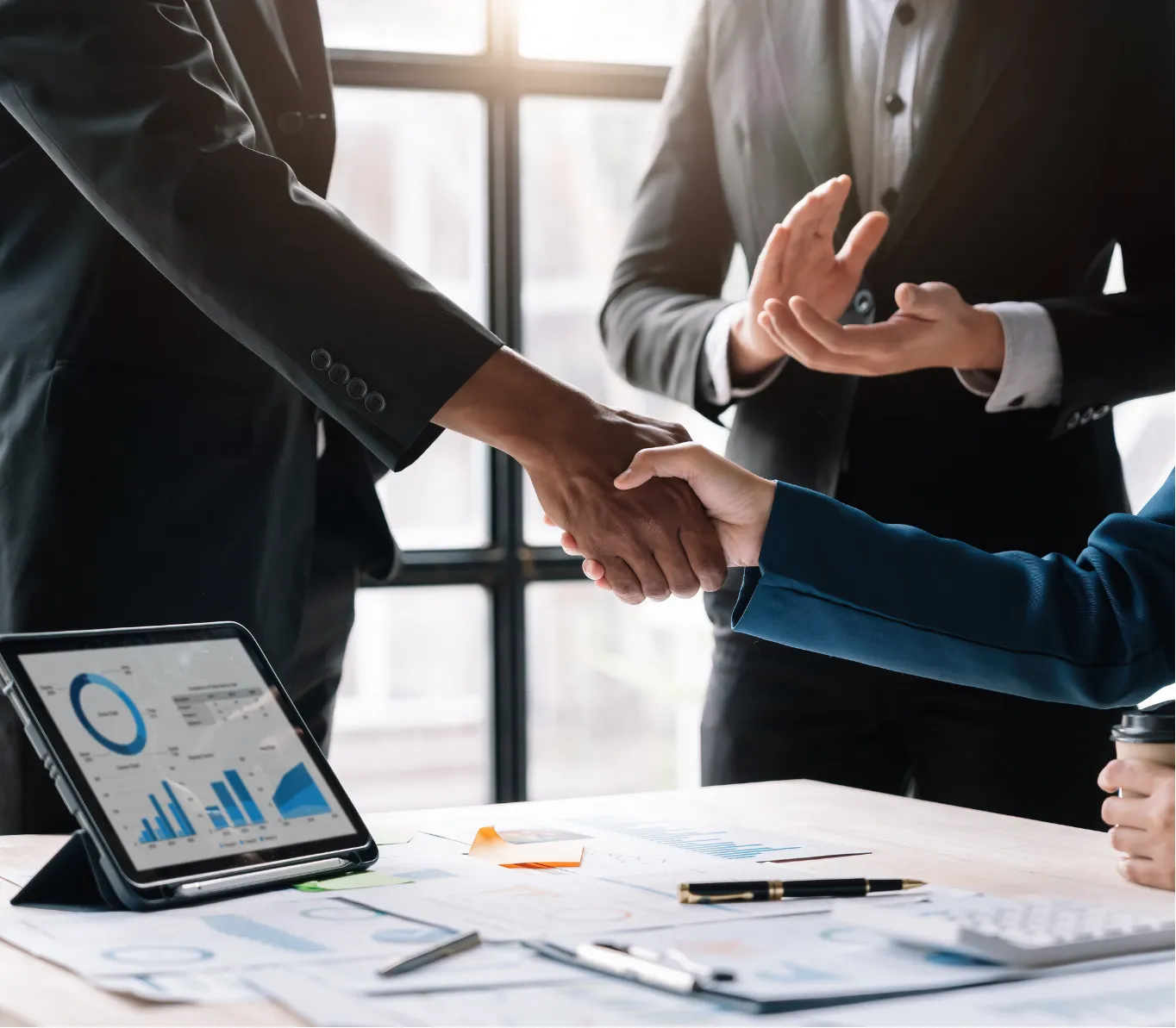 mergers-2 Two people in business attire shaking hands over a desk with charts and a tablet, while another person gestures.