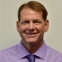 Man in a black suit and lavender tie smiling in front of a light gray background.