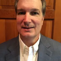 Man in a black suit and lavender tie smiling in front of a light gray background.