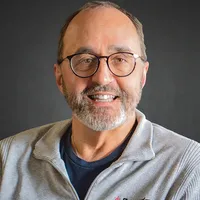 Man in a black suit and lavender tie smiling in front of a light gray background.