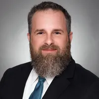 Man in a black suit and lavender tie smiling in front of a light gray background.