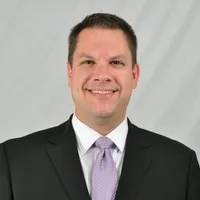Man in a black suit and lavender tie smiling in front of a light gray background.