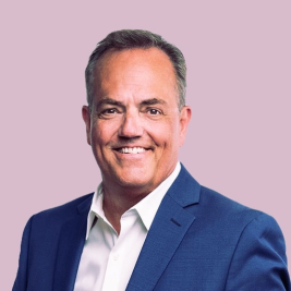 Man in a black suit and lavender tie smiling in front of a light gray background.