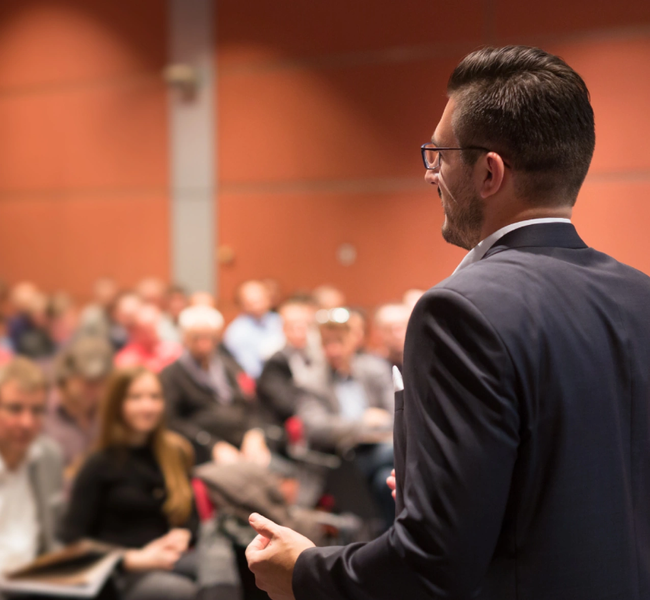 A man in a suit speaks to an audience in a conference room, with people seated and listening attentively.